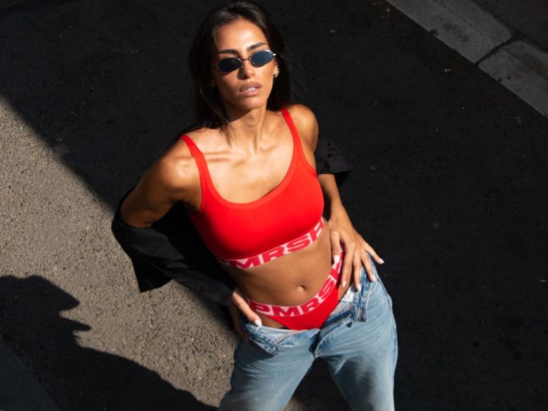 Devojka pozira na ulici, na njoj su nabolje tange i top u crvenoj boji kao i košulja, naočare i farmerke