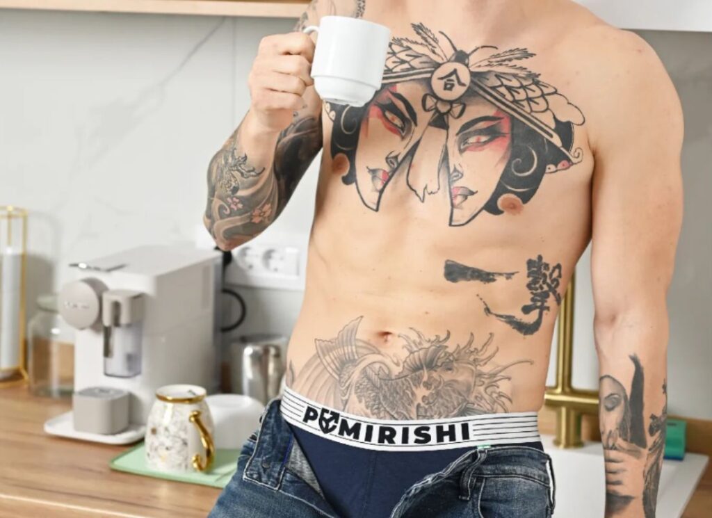 momak sa otkopčanim pantalonama stoji naslonjen na kuhinjsku policu i drži šolju kafe u ruci